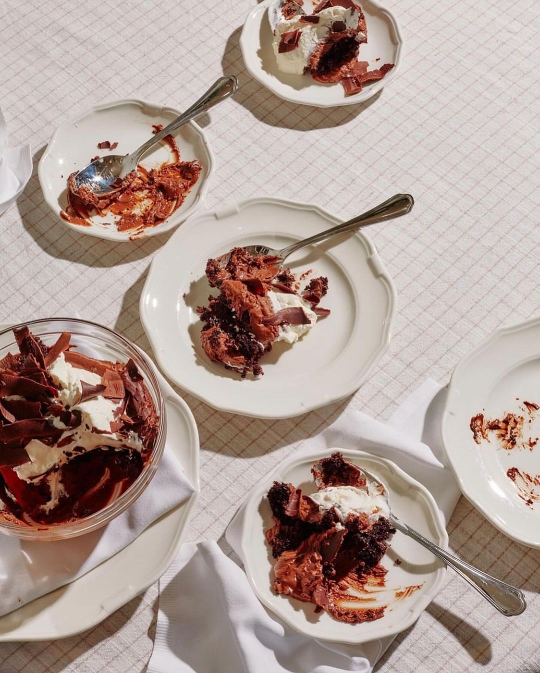 Overhead shot of individual dessert plates with spoons and chocolate dessert with whipped cream on a white tablecloth in warm lighting.