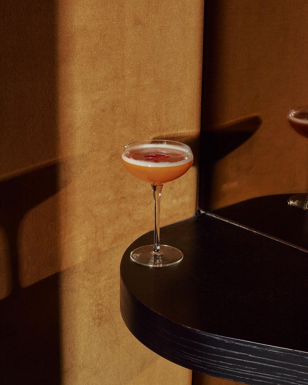 A coupe glass filled with a reddish-brown cocktail sits on a black table against a textured brown wall, side lit.