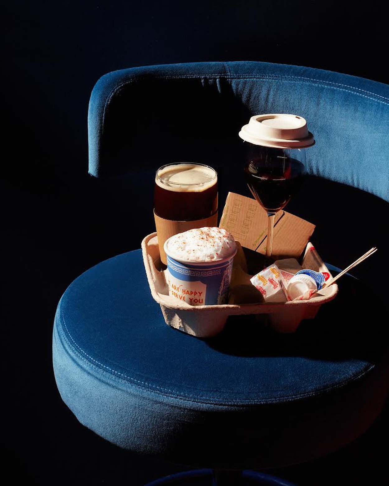 Coffee and wine in a paper carrier on a blue velvet chair, set against a dark background, creating a cozy and inviting atmosphere.