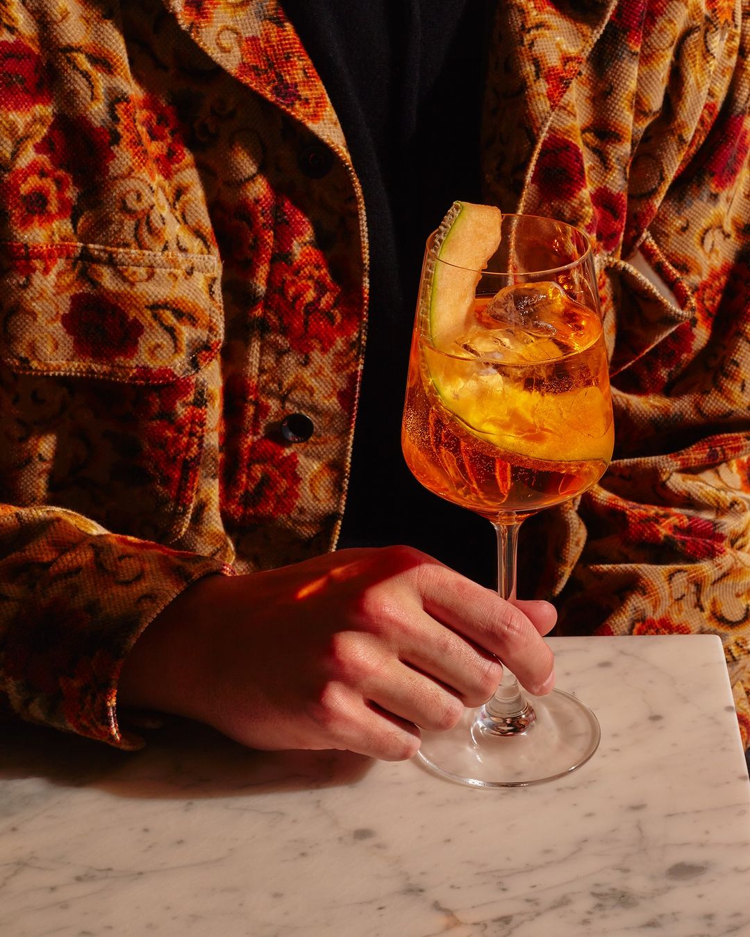 Person in floral jacket holds an aperitivo cocktail in a stemmed glass with a melon garnish, set on a marble counter in a warm light.