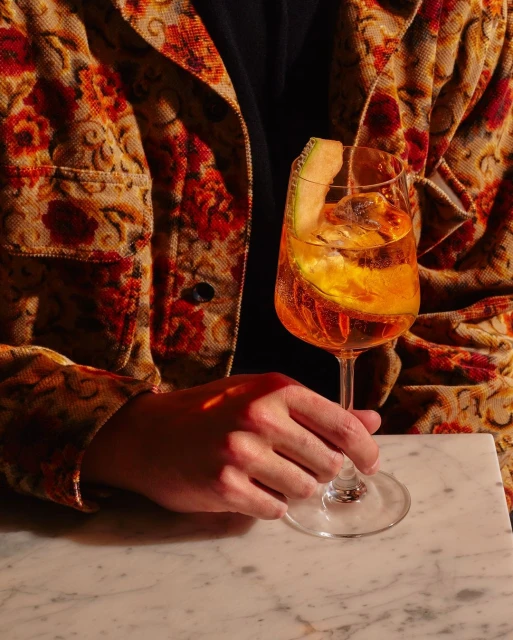 Person in floral jacket holds an aperitivo cocktail in a stemmed glass with a melon garnish, set on a marble counter in a warm light.