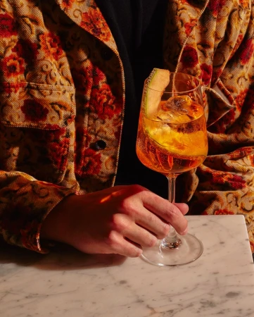 Person in floral jacket holds an aperitivo cocktail in a stemmed glass with a melon garnish, set on a marble counter in a warm light.