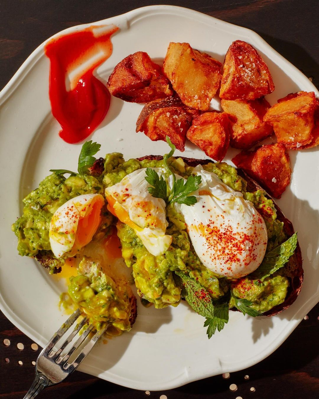 Overhead shot of avocado toast with poached eggs, roasted potatoes, and ketchup on a white plate, showcasing a vibrant food presentation.