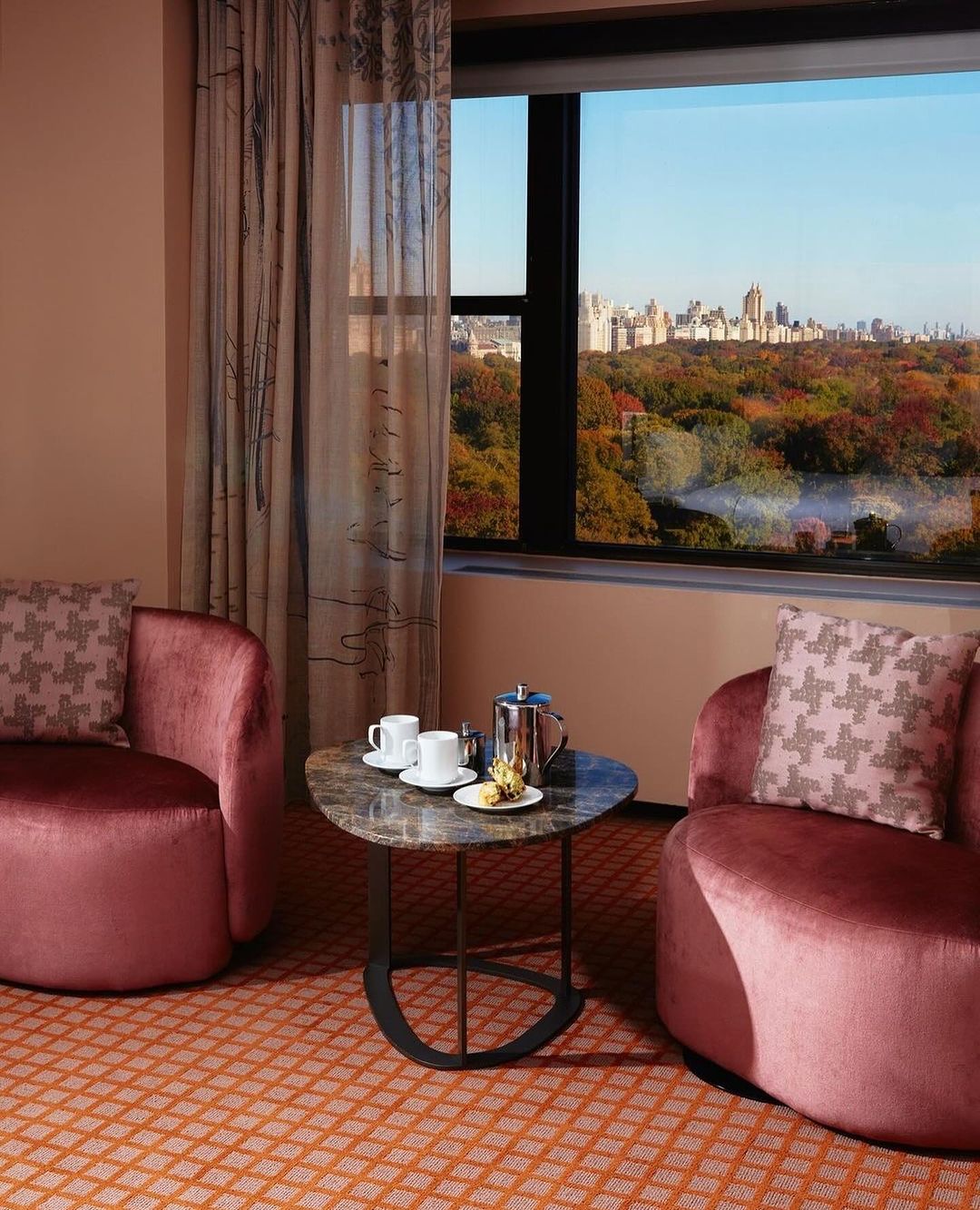 A comfortable hotel room scene with modern armchairs, a round marble table set for tea, and a window view of a cityscape and park.