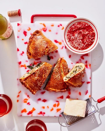 Overhead shot of sliced calzones filled with spinach and cheese, served with tomato sauce and grated cheese, on a patterned cutting board.