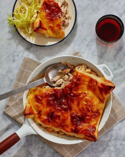 Overhead shot of chicken pot pie in a white skillet with flaky crust, a serving of greens, and a drink on a marble surface.