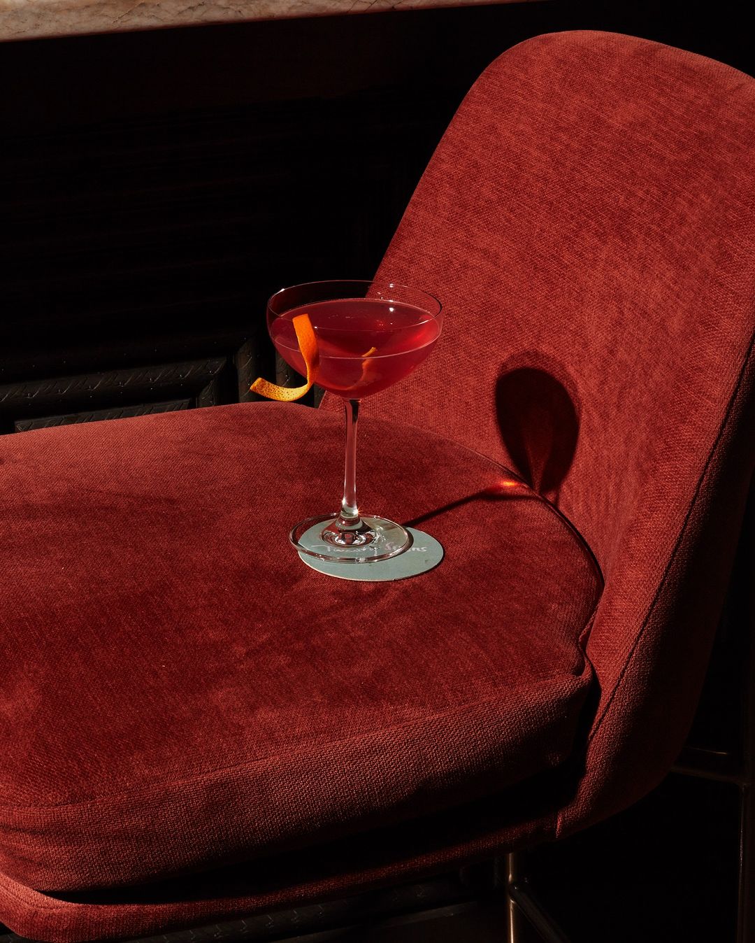 A red cocktail in a coupe glass with an orange peel garnish sits on a red velvet chair in a dimly lit bar setting.