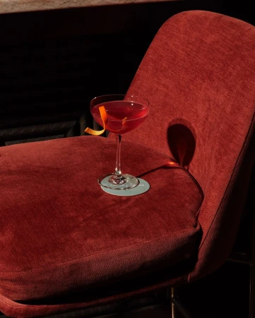 A red cocktail in a coupe glass with an orange peel garnish sits on a red velvet chair in a dimly lit bar setting.