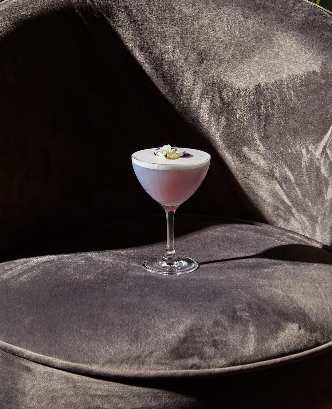 Elegant presentation of a light-purple cocktail garnished with edible flowers, sitting on a velvet chair in dramatic chiaroscuro lighting.