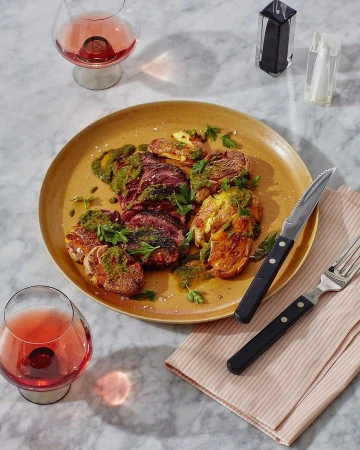 Overhead shot of smashed potatoes on a plate with green sauce, accompanied by silverware and glasses of rose wine on a marble surface.