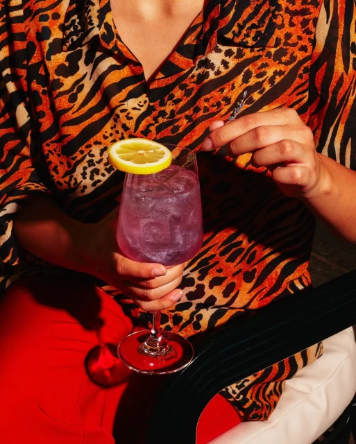 Person in tiger-print shirt holds sparkling purple cocktail with lemon slice garnish, seated indoors with bright red pants.