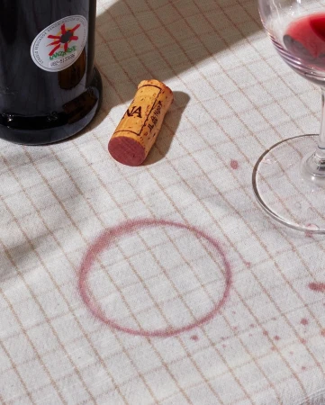 Still life of a wine bottle, cork, glass, and a wine stain on a patterned tablecloth in natural light, capturing a rustic and candid scene.
