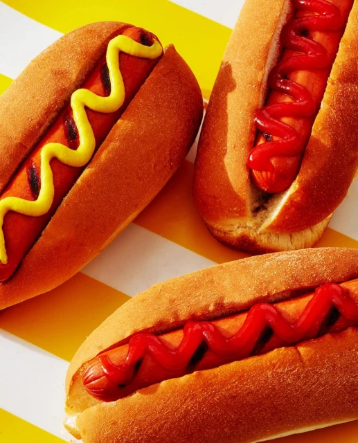 Three hot dogs with mustard and ketchup on a striped background. The hot dogs are in focus with a simple composition.