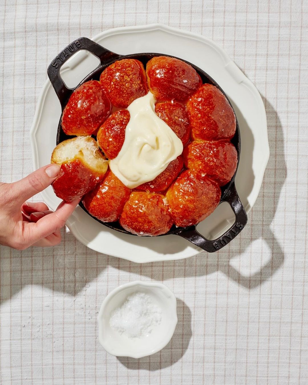 Overhead shot of black cast iron skillet filled with glistening monkey bread topped with cream, with hand taking one piece.