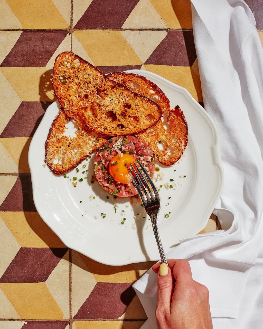 Overhead shot of beef tartare topped with an egg yolk on a plate with toast points, garnished with herbs, on a patterned tile surface.