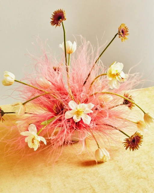 Floral arrangement featuring white daffodils, tulips, and gerbera daisies with pink decorative grass in a white vase on a yellow surface.