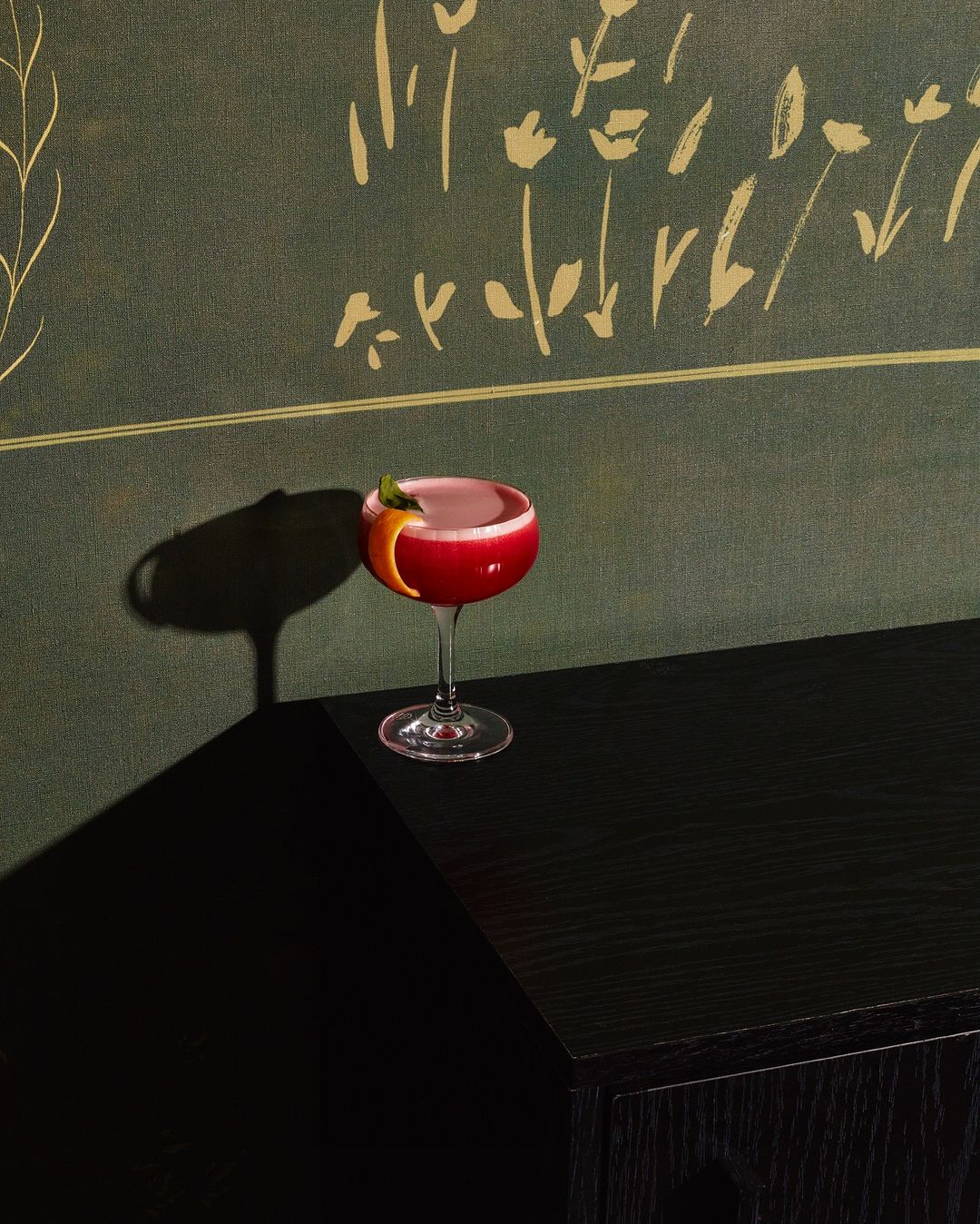 Elegant cocktail in coupe glass on black cabinet, set against green botanical wallpaper, casting strong shadow in dark room.