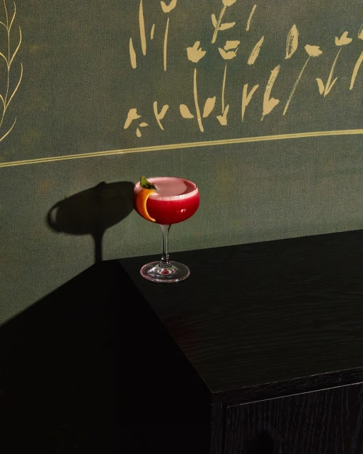 Elegant cocktail in coupe glass on black cabinet, set against green botanical wallpaper, casting strong shadow in dark room.