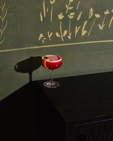 Elegant cocktail in coupe glass on black cabinet, set against green botanical wallpaper, casting strong shadow in dark room.