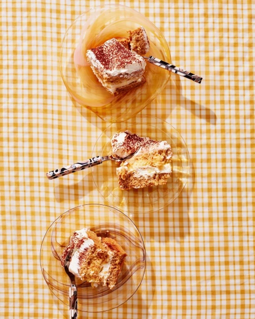 Overhead shot of three slices of tiramisu on clear glass plates, each with a spoon, on a yellow and white checkered tablecloth.