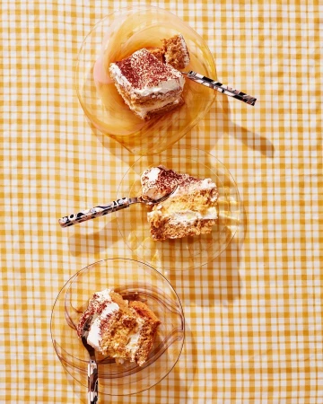 Overhead shot of three slices of tiramisu on clear glass plates, each with a spoon, on a yellow and white checkered tablecloth.
