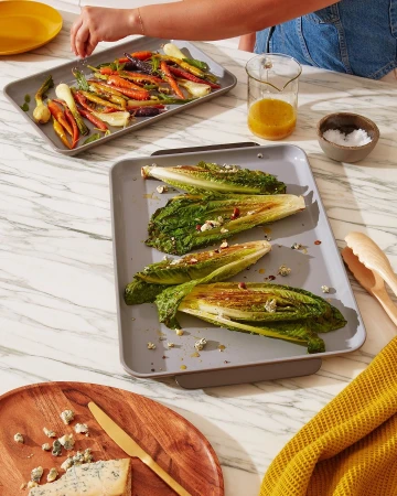 Overhead shot features roasted romaine lettuce hearts, rainbow carrots, and culinary plating atop a marble surface in warm lighting.