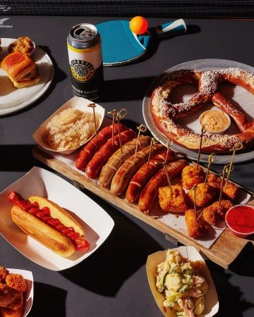 Overhead shot of hotdogs, sausages, pretzels, and beer on a ping pong table, highlighting food photography and table tennis.