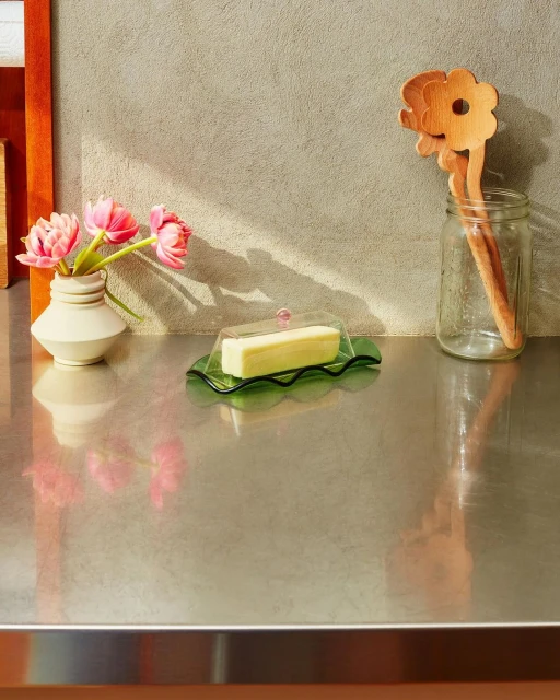 Still life of butter dish, flower vase, and utensils on a stainless steel counter with soft, natural lighting and muted background.