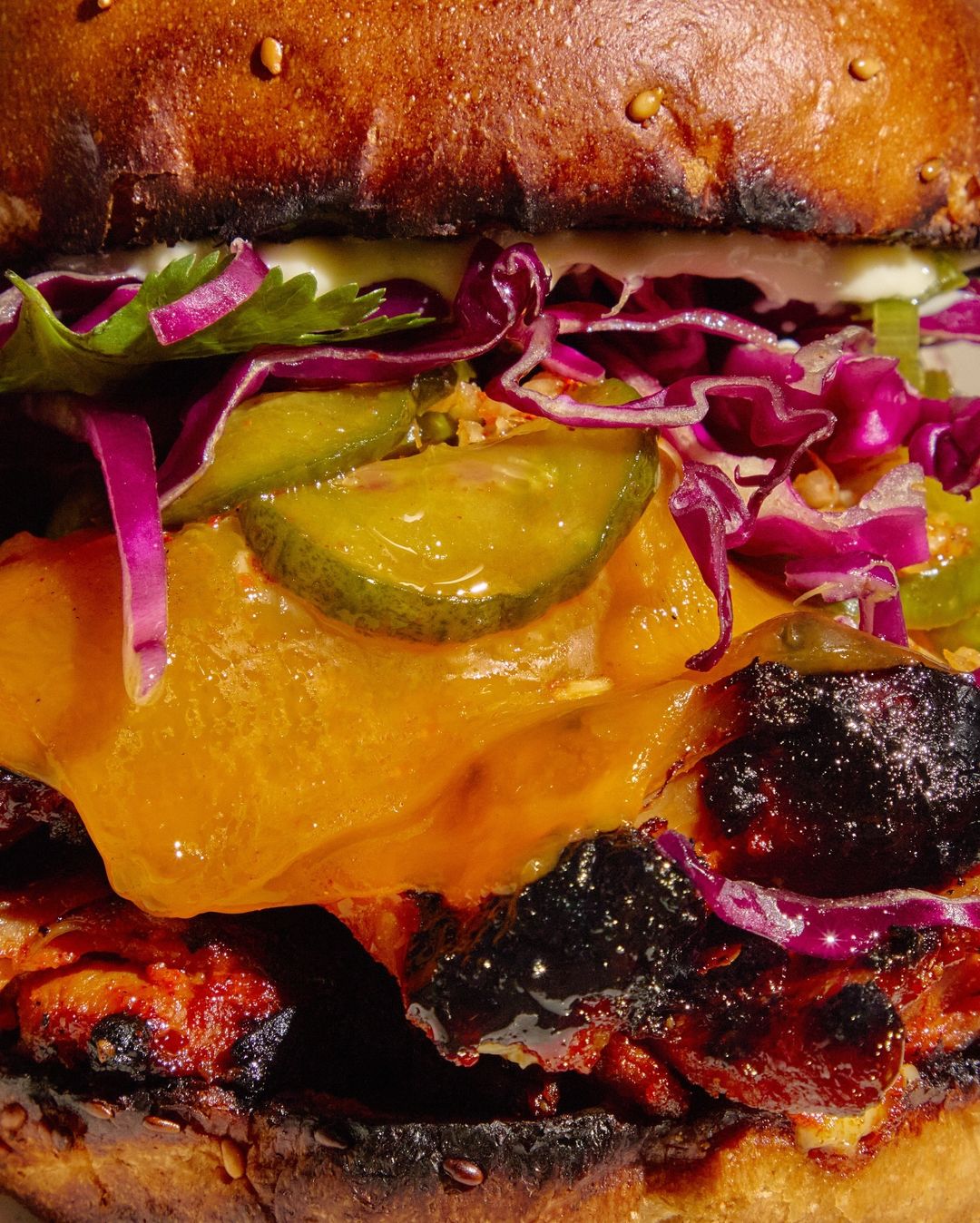 Close-up of a gourmet burger featuring layers of charred meat, melted cheese, crisp pickles, and red cabbage, topped with a glazed bun.