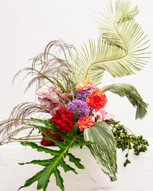 Artistic floral arrangement featuring orange and purple flowers, green foliage, and tall brown grasses against a white backdrop.