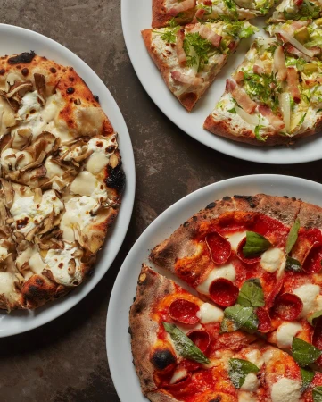 Overhead shot of three pizzas with different toppings on white plates and a dark surface, displaying culinary variety.