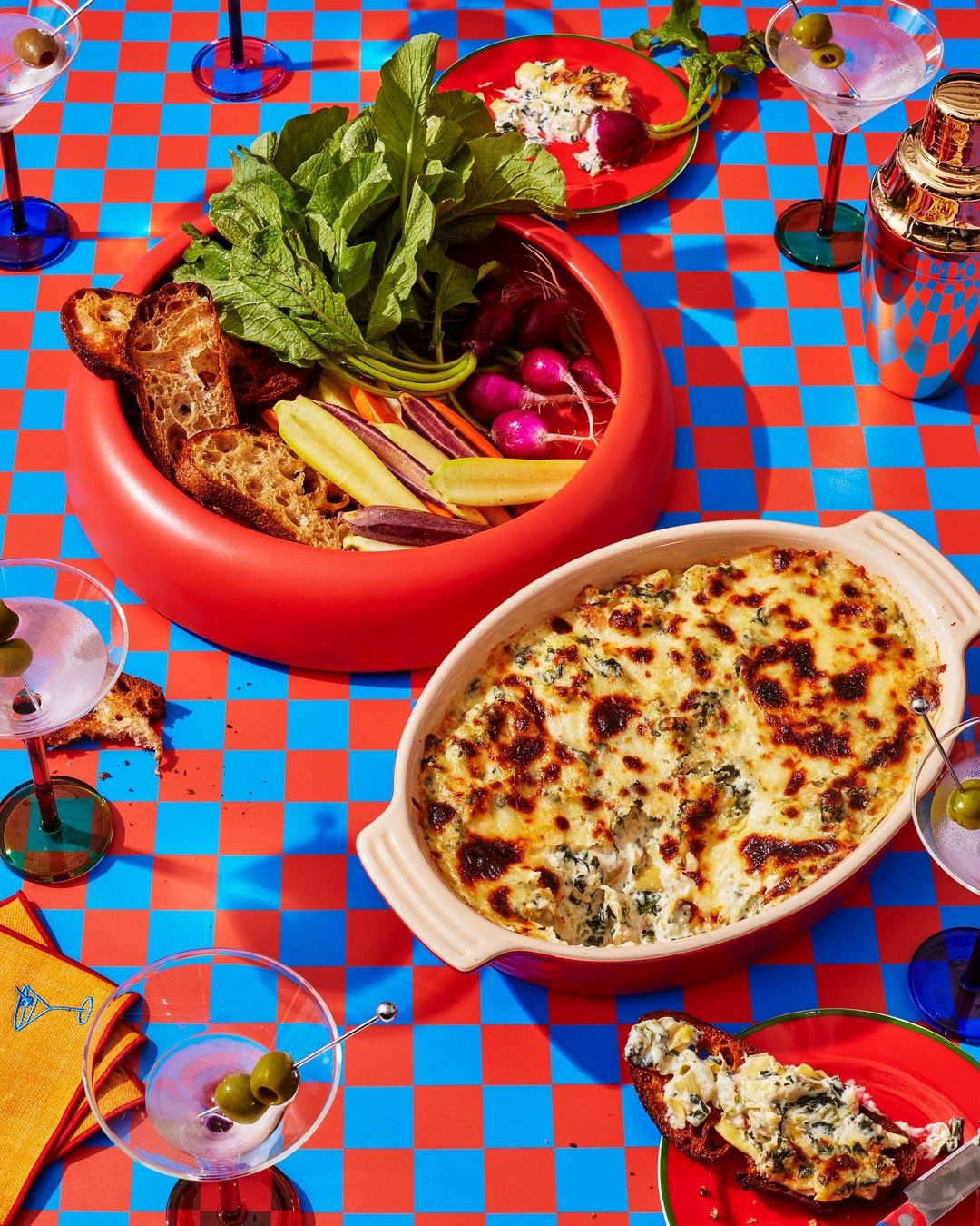 Overhead shot of a vibrant checkerboard table laden with appetizers: a veggie platter, cheesy dip, cocktails, and crostini.