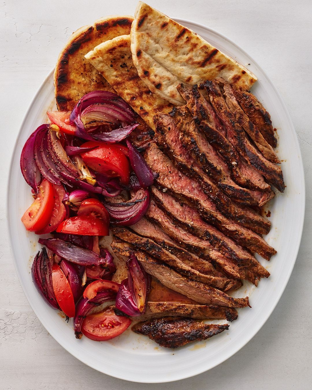 Grilled steak slices, sliced tomatoes and red onions, and pita bread on an oval white plate, presenting a delectable food arrangement.