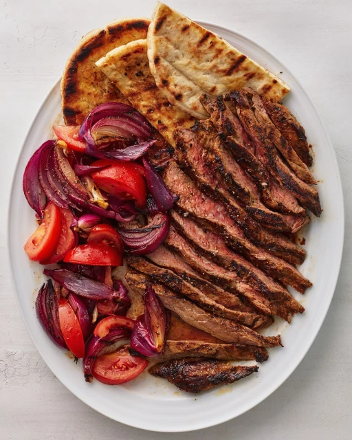 Grilled steak slices, sliced tomatoes and red onions, and pita bread on an oval white plate, presenting a delectable food arrangement.