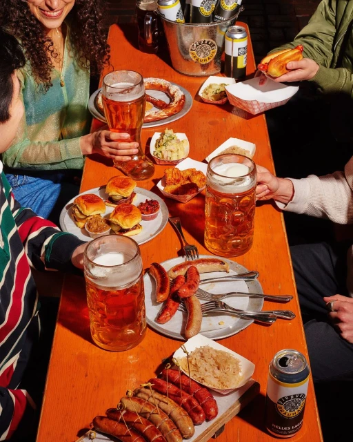 Overhead shot of four individuals gathered around a table filled with beer, sausages, and pretzels in an indoor setting. Food and drink.