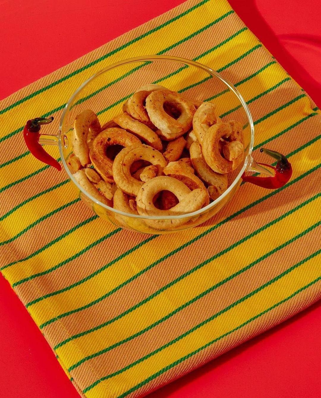 A glass bowl filled with taralli crackers sits on a striped yellow and orange cloth against a bright red background, food still life.