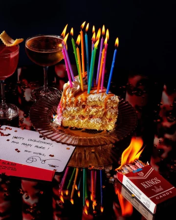 Celebratory still life shot on a reflective surface, featuring a slice of cake with candles, cocktails, and a burning pack of cigarettes.