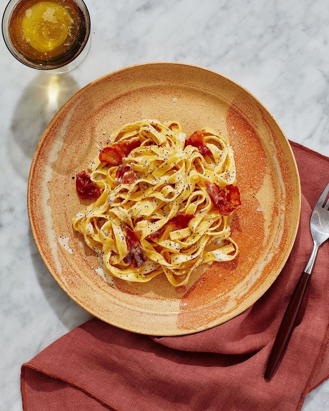 Overhead shot of pasta carbonara on a ceramic plate with a fork and a glass of white wine. Table with linen napkin.