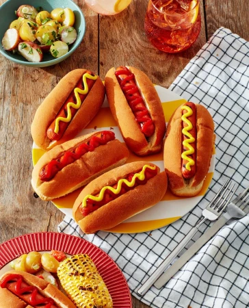 Overhead shot of hot dogs in buns with mustard and ketchup, side dishes, and drinks on a wooden picnic table with a checkered cloth.