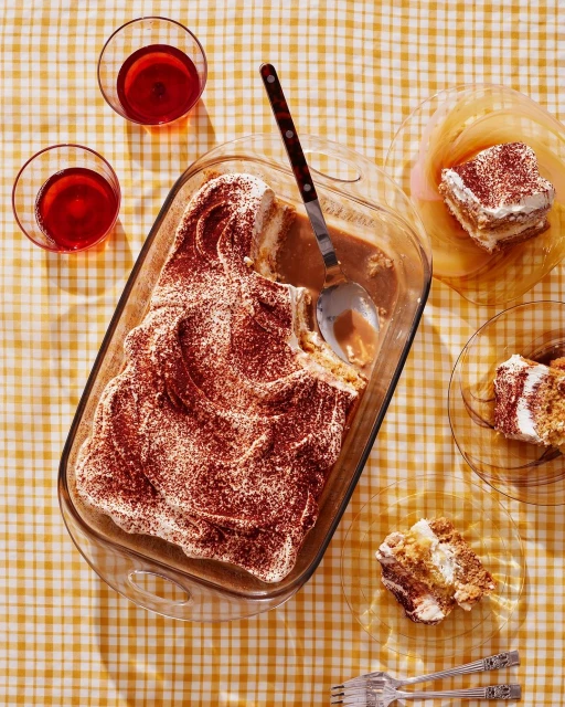 Overhead shot of a tiramisu dessert in a glass baking dish, served with dessert plates, forks, and glasses of red liquid on a table.