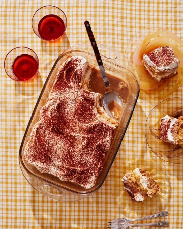 Overhead shot of a tiramisu dessert in a glass baking dish, served with dessert plates, forks, and glasses of red liquid on a table.