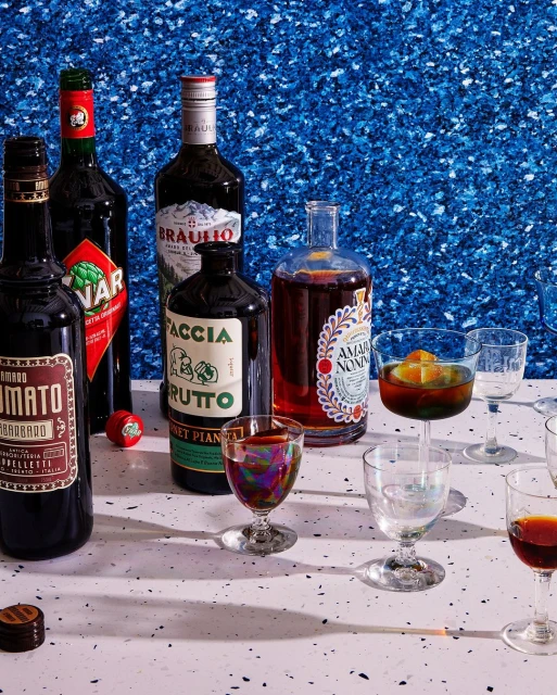 Still life of assorted amaro bottles and stemmed cocktail glasses arranged on a speckled surface against a blue textured background.