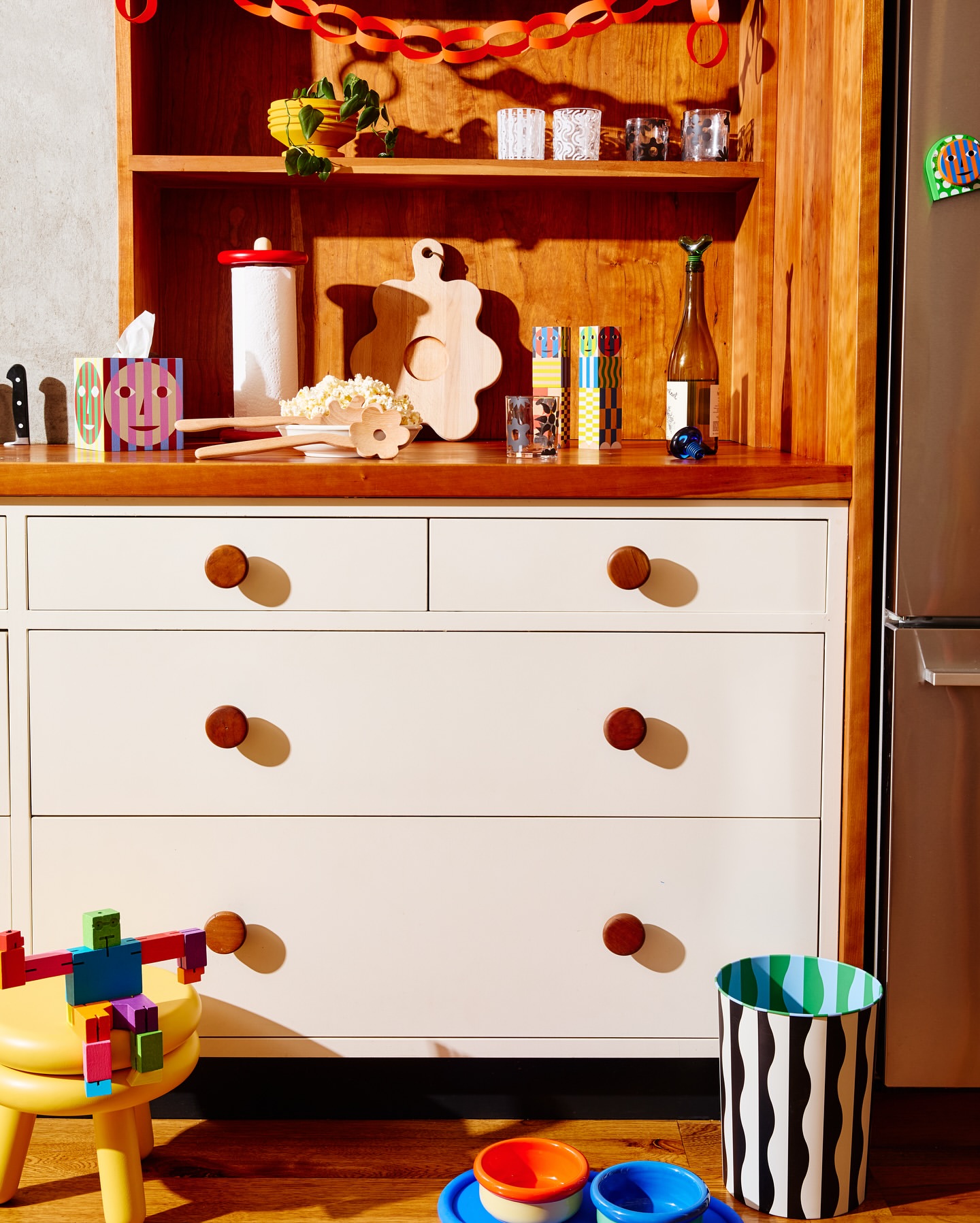 Interior shot of a white dresser with wooden knobs, a yellow stool, and colorful objects in a warmly lit room, creating a cozy atmosphere.