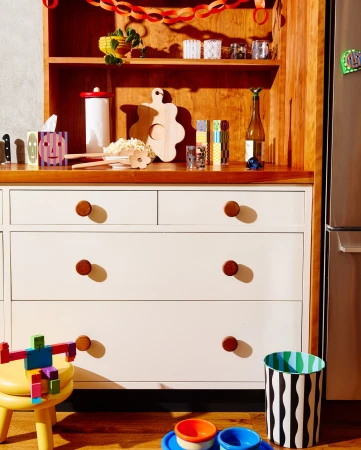 Interior shot of a white dresser with wooden knobs, a yellow stool, and colorful objects in a warmly lit room, creating a cozy atmosphere.