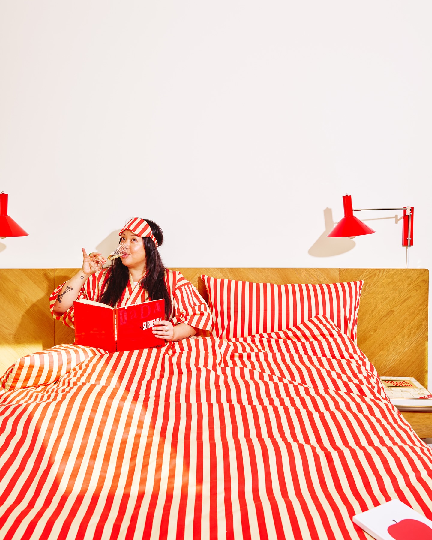 Woman in striped pajamas relaxes in bed with red book, sipping wine in a brightly lit room with red and white striped bedding.
