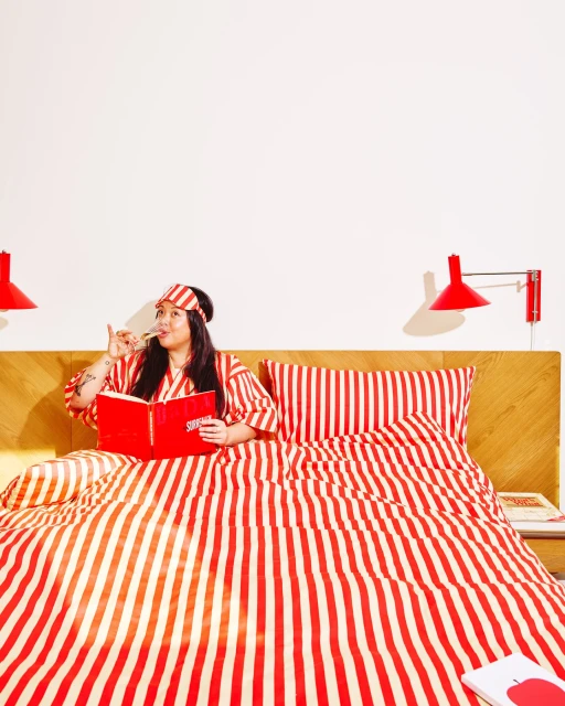 Woman in striped pajamas relaxes in bed with red book, sipping wine in a brightly lit room with red and white striped bedding.