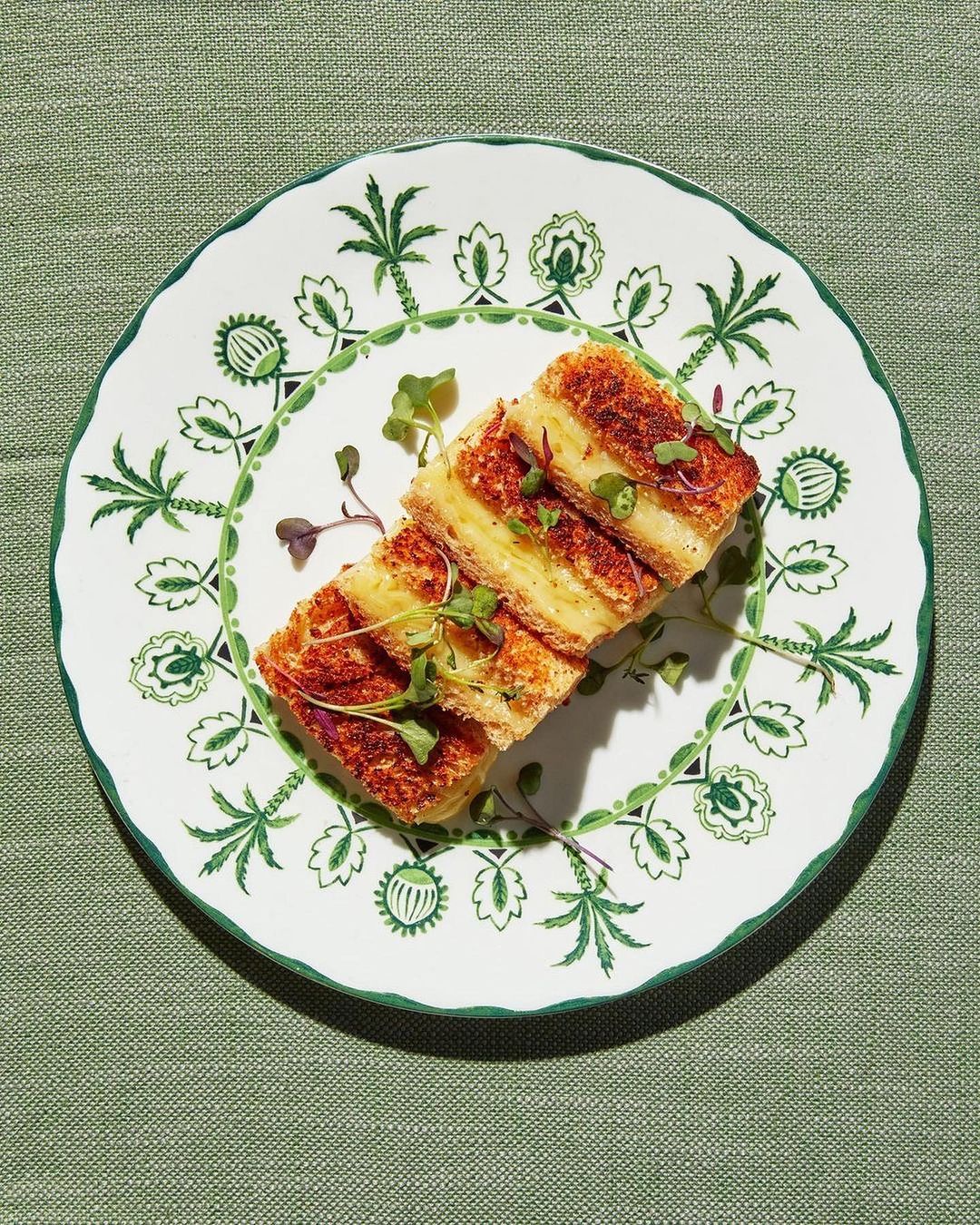 Grilled cheese sandwich cut into sections on a decorative plate with garnish, set on a green textured surface.