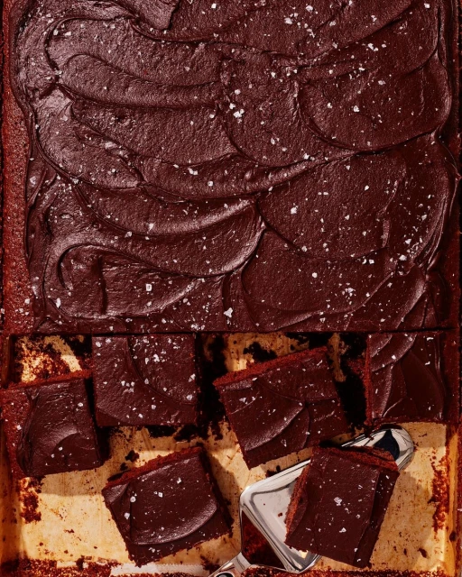 Close-up of a sheet pan of freshly baked, dark chocolate cake with a rich chocolate frosting, cut into small squares and sprinkled with salt.