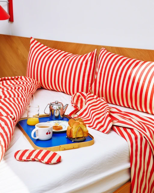 Overhead shot of breakfast in bed: croissants, coffee, orange juice and sleeping mask arranged on blue tray with red striped bedding.