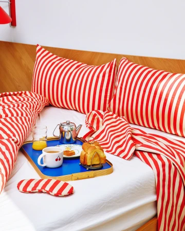 Overhead shot of breakfast in bed: croissants, coffee, orange juice and sleeping mask arranged on blue tray with red striped bedding.